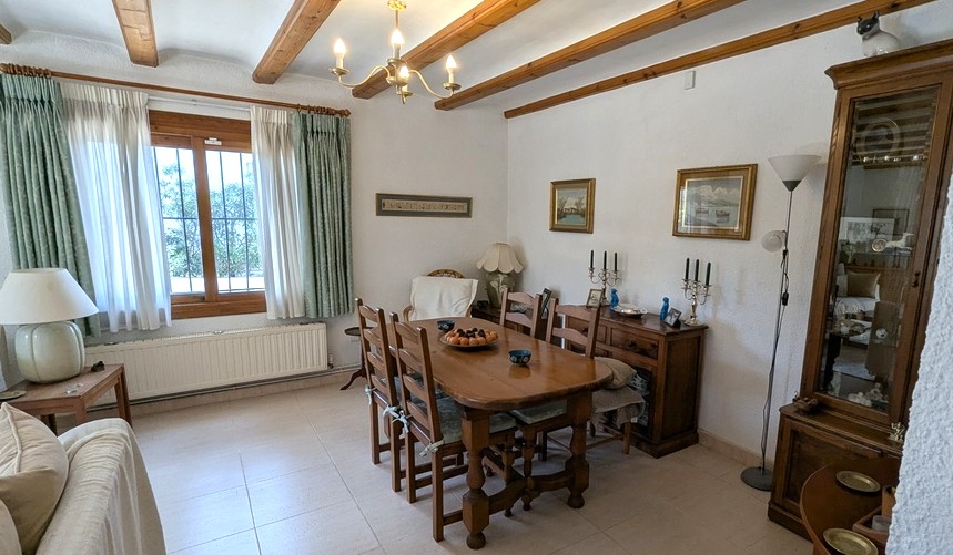 Traditioneller Essbereich mit sichtbaren Holzbalken, Massivholztisch und klassischer Anrichte. Der Raum verfügt über einen Heizkörper und Fenster mit Sicherheitsgittern.