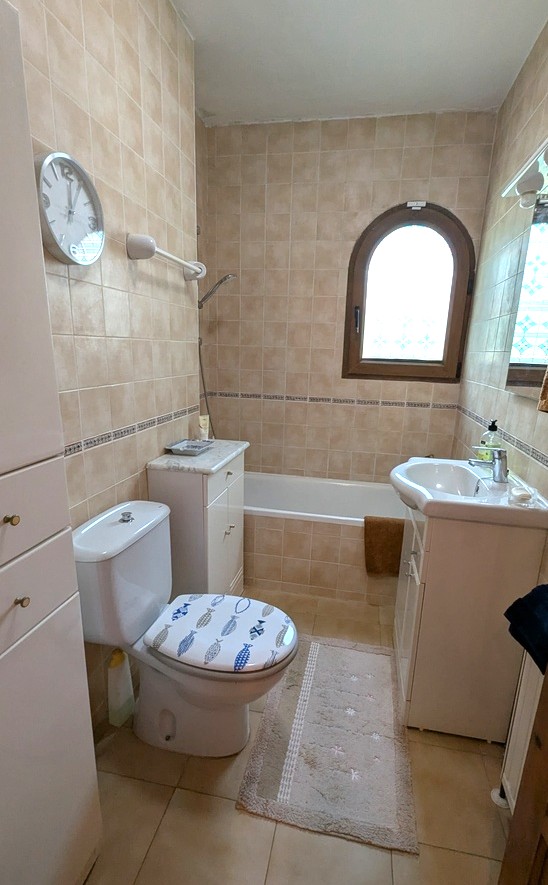 Traditionelles Badezimmer mit beigen Wandfliesen, Einbaubadewanne und einem Rundbogenfenster. Ausgestattet mit weißem Waschtisch und praktischen Aufbewahrungsschränken.
