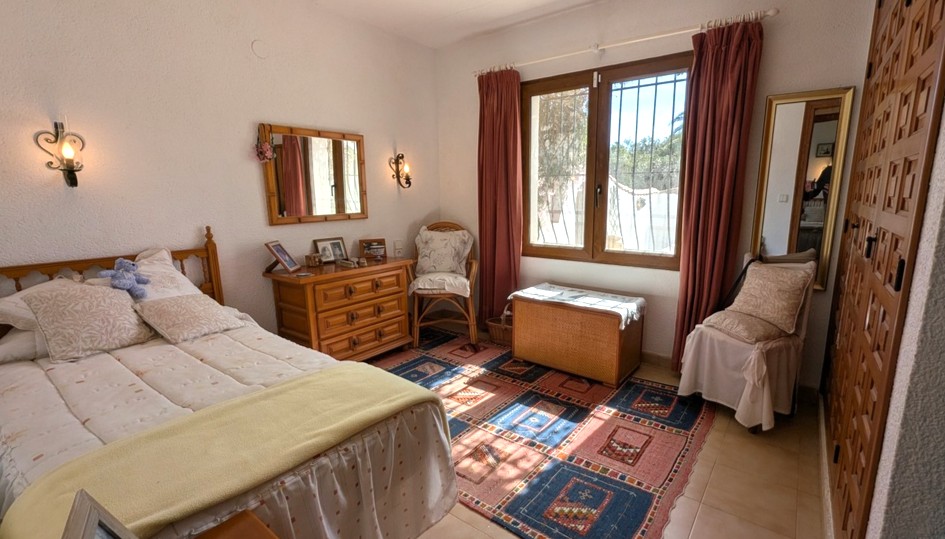 Traditionelles Schlafzimmer mit Einbauschränken aus Holz, passender Kommode und Tageslicht. Strukturierte Wände und Fliesenboden mit gemustertem Teppich.
