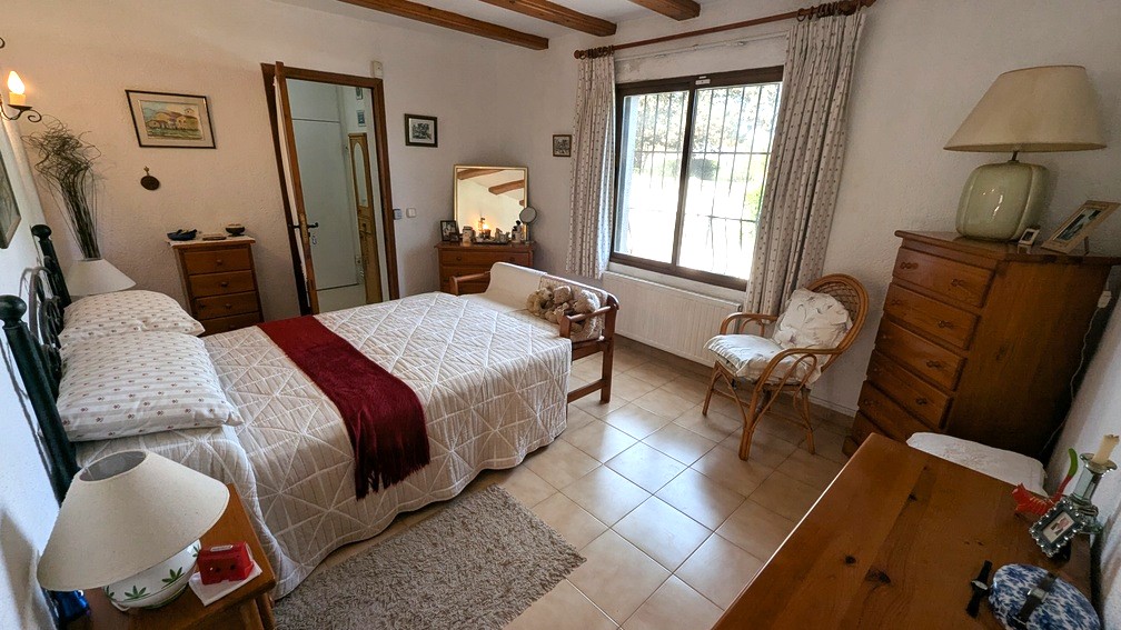 Traditionelles Schlafzimmer mit Holzbalkendecke, Fliesenboden und Heizkörper unter dem Fenster, das viel Tageslicht in den Raum lässt.
