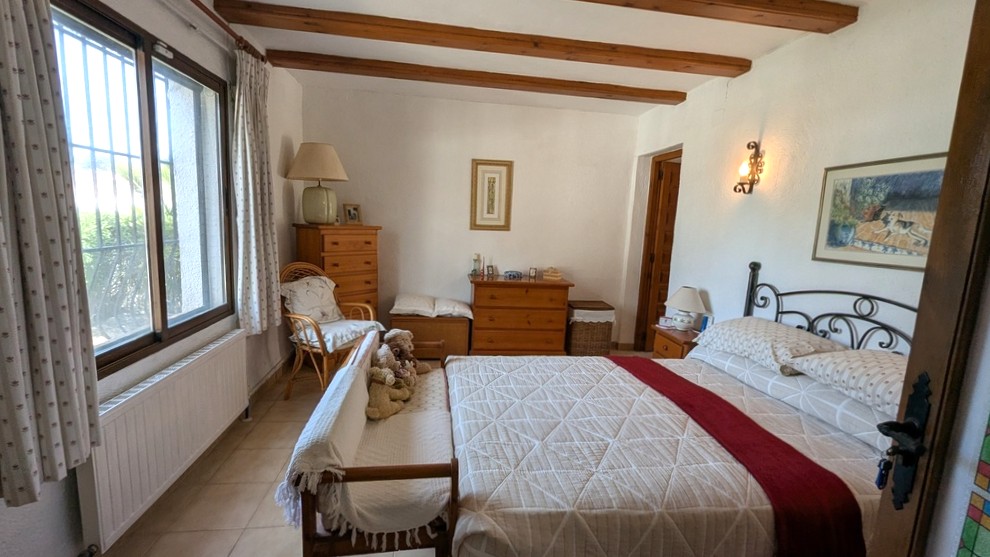 Traditionelles Schlafzimmer mit sichtbaren Holzbalken, einem schmiedeeisernen Bettgestell und klassischen Holzmöbeln. Viel Tageslicht sorgt für eine helle Atmosphäre.