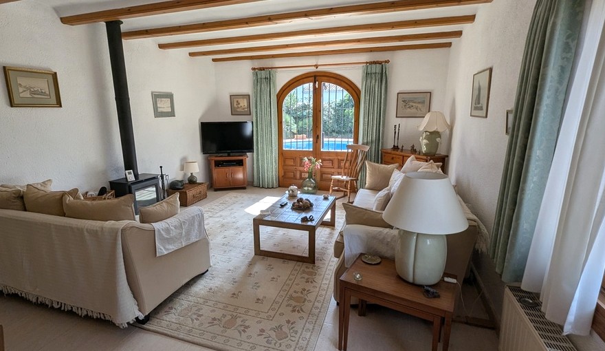 Wohnzimmer mit sichtbaren Holzbalken, Kaminofen und bogenförmigen Holztüren zum Poolbereich. Traditionelle Möbel und neutrale Farbtöne unterstreichen den rustikalen Stil.