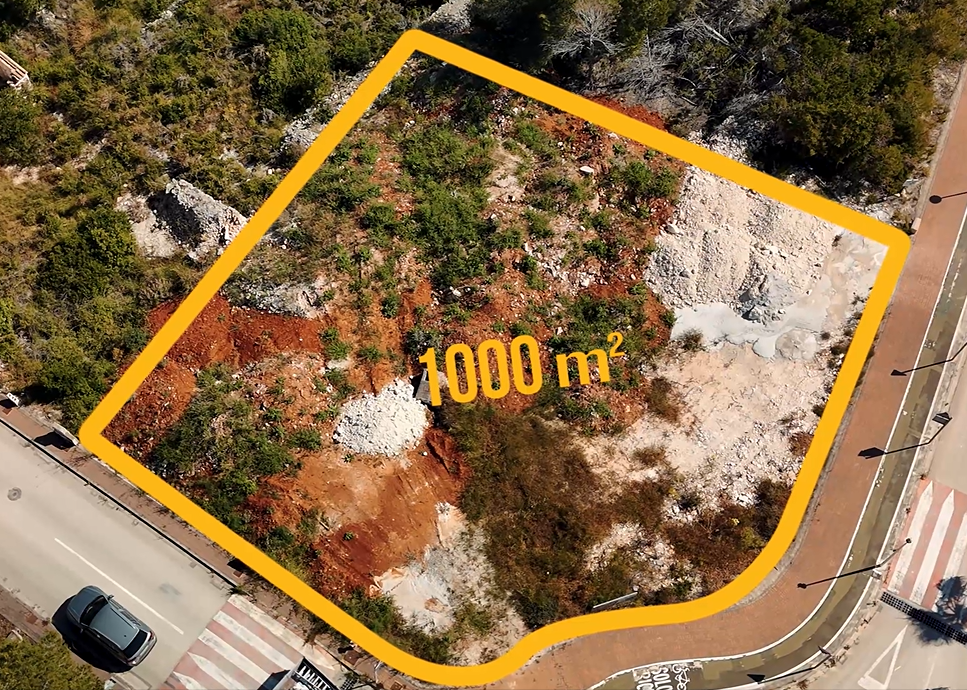 Luftaufnahme des 1.000 m² großen Eckgrundstücks in Jávea mit rötlichem Boden, natürlicher Vegetation und voll erschlossener Straßenanbindung samt Gehwegen.