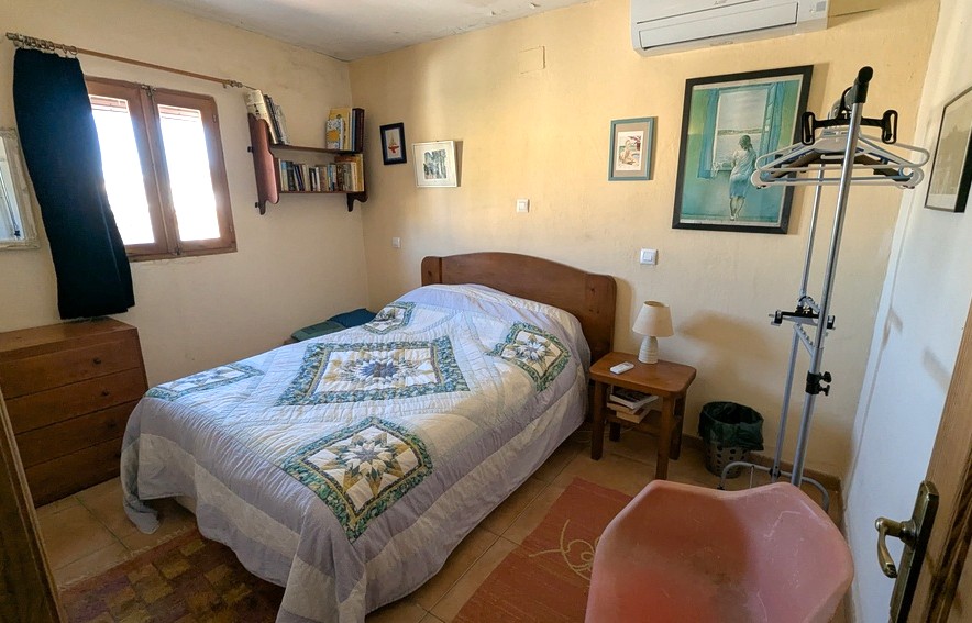 Schlafzimmer mit Holzmöbeln, Fliesenboden und Split-Klimaanlage. Verfügt über Wandregale, eine klassische Kommode und natürliches Licht durch ein Holzfenster.