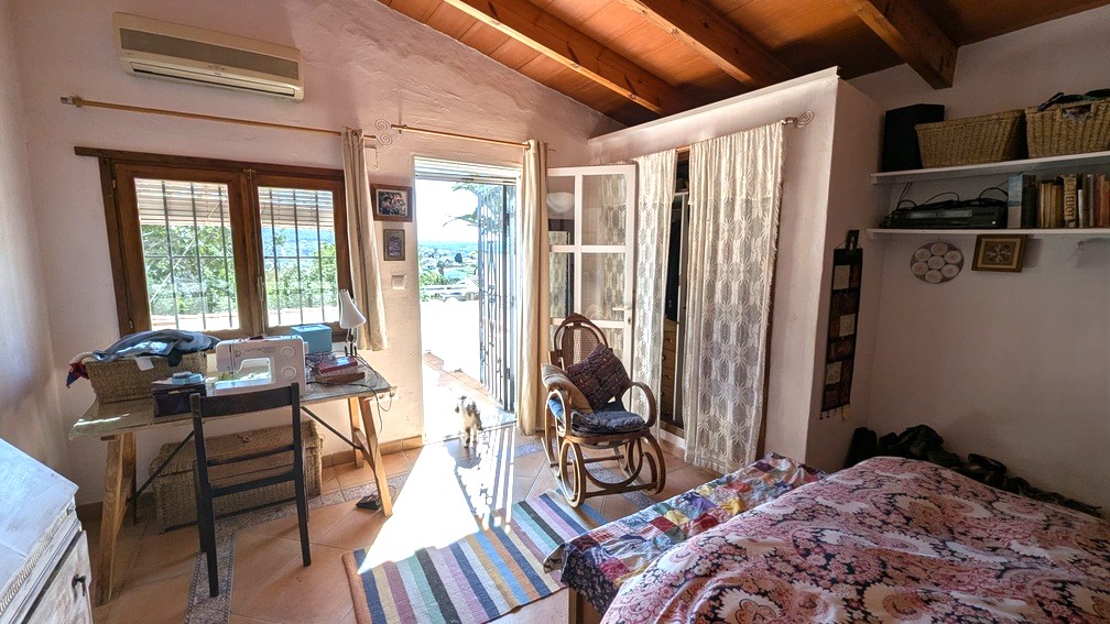 Schlafzimmer mit sichtbaren Holzbalken, Terrakottaboden und direktem Zugang zur Terrasse mit Blick ins Grüne. Ausgestattet mit Klimaanlage und Arbeitsbereich.