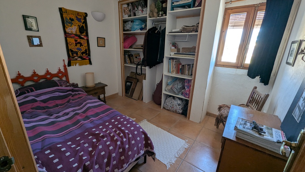 Schlafzimmer mit Terrakotta-Fliesenboden, integrierten offenen Regalen und Holzfensterrahmen. Der Raum bietet ein Einzelbett und einen Schreibtisch bei natürlichem Licht.