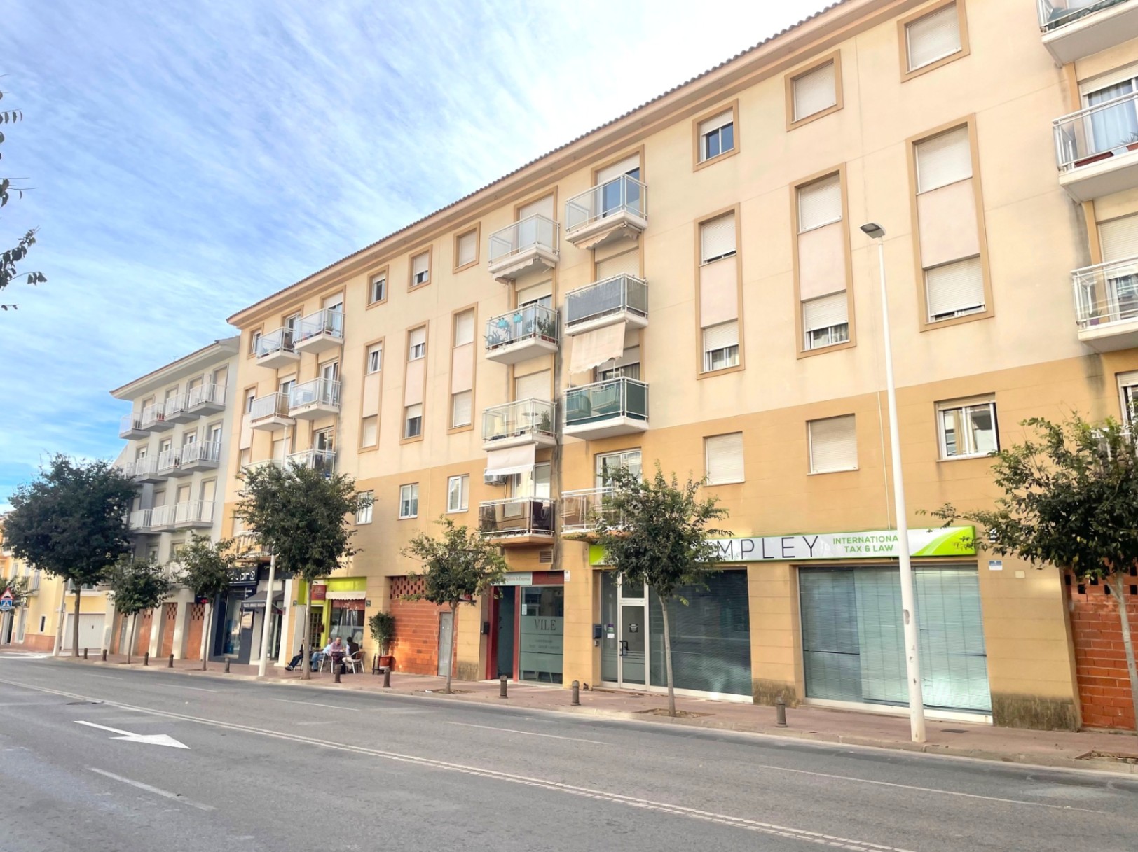 Außenfassade eines mehrstöckigen Wohngebäudes mit Gewerbeeinheiten im Erdgeschoss an einer breiten Allee mit Bäumen und modernen Glasbalkonen.