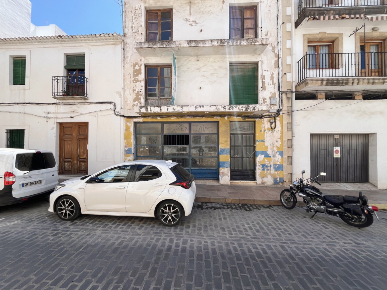 Außenfassade des Gebäudes in Jávea mit Gewerbefläche im Erdgeschoss, Balkonen in den oberen Etagen und Holzfenstern an einer traditionellen Kopfsteinpflasterstraße.