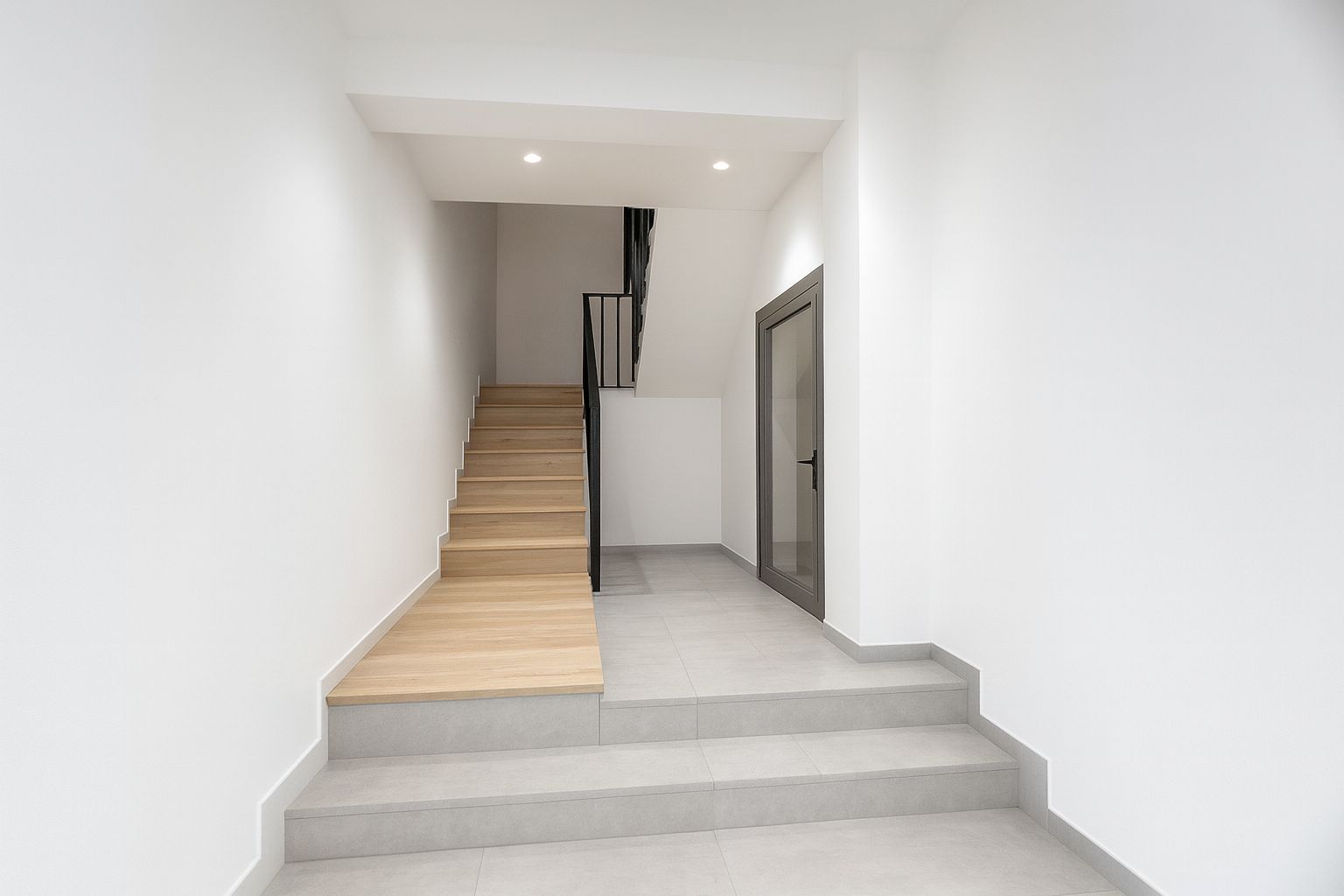 Eingangsbereich mit einer Treppe aus hellem Holz und schwarzem Metallgeländer. Graue Bodenfliesen, weiße Wände und eine Glastür mit grauem Rahmen.