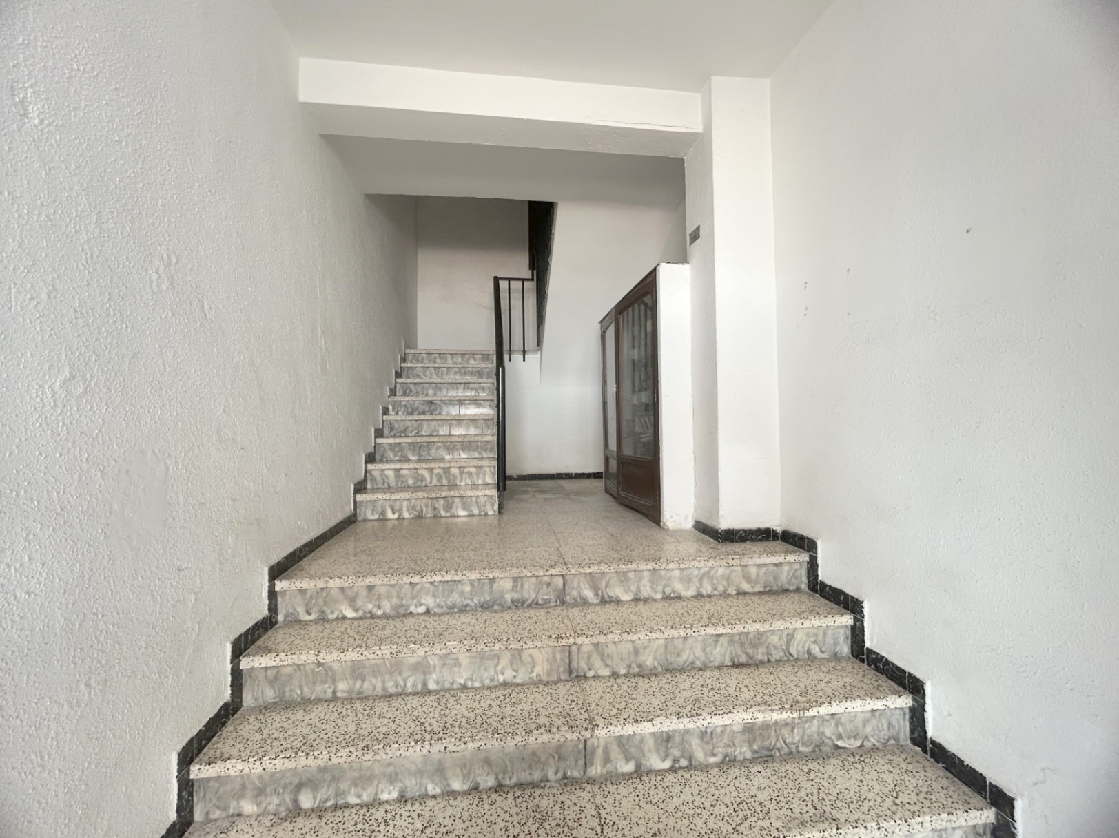 Gemeinschaftstreppenhaus mit Terrazzostufen, marmorierten Kanten und schwarzem Metallgeländer. Ein Holzschrank auf dem Zwischenpodest ergänzt den funktionalen Eingangsbereich.