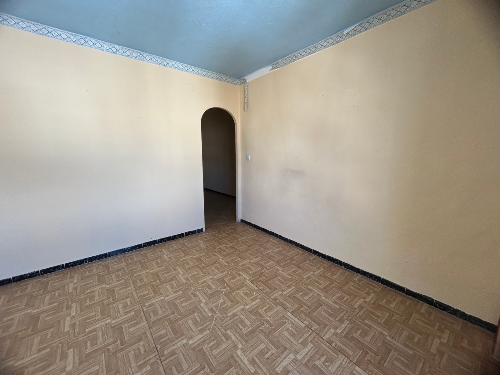 Innenraum mit gemusterten Keramikfliesen, cremefarbenen Wänden und einer hellblauen Decke mit Zierborte. Ein Rundbogen führt in den angrenzenden Bereich.