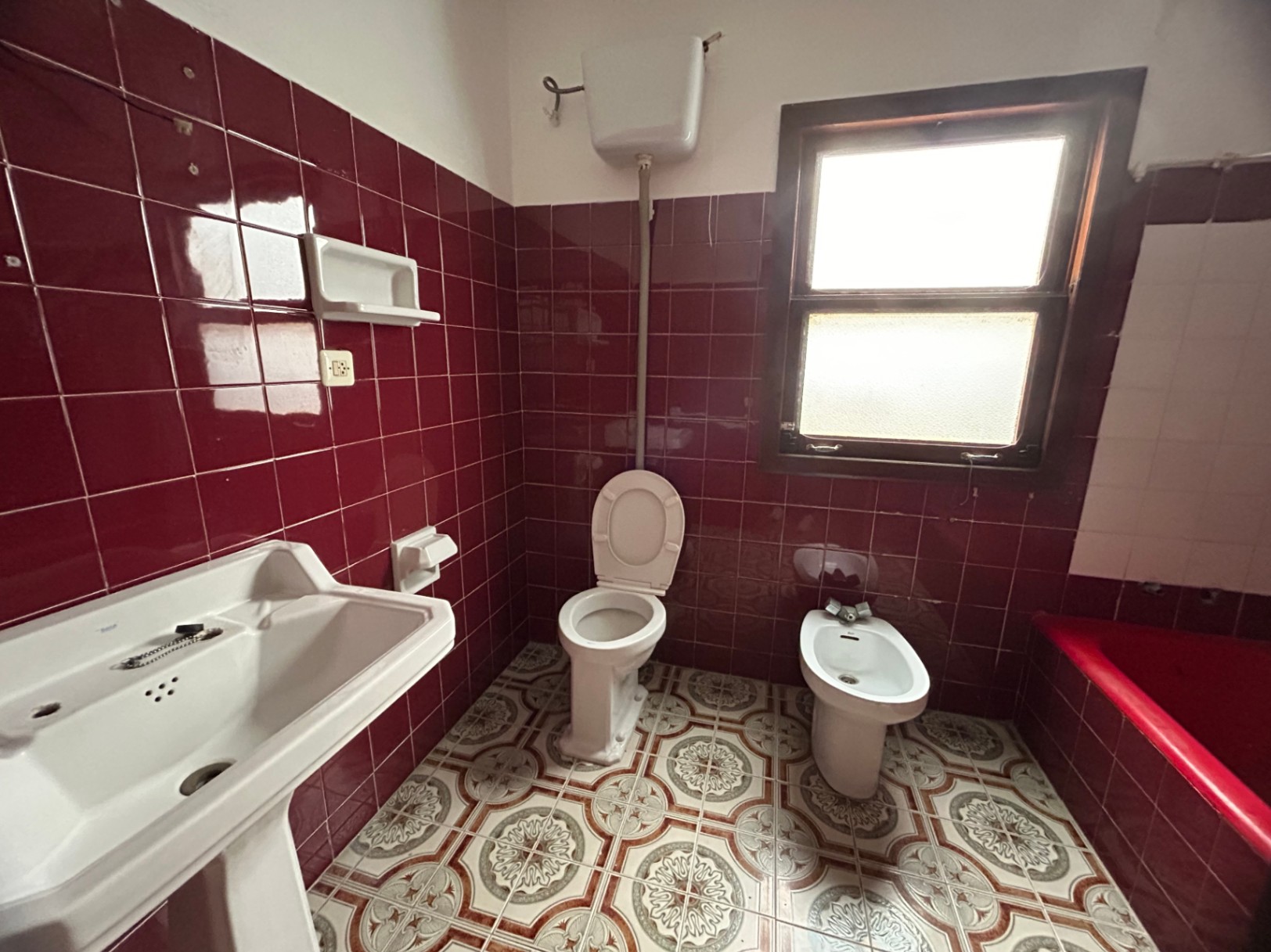 Originales Badezimmer mit weinroten Wandfliesen, gemustertem hydraulischem Boden und klassischen Sanitärobjekten. Ein Holzfenster sorgt für natürliches Licht und Belüftung.