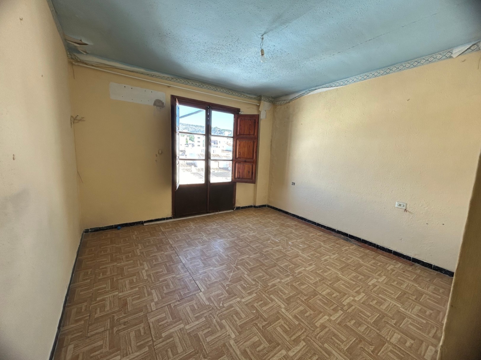 Renovierungsbedürftiger Raum mit gemusterten Keramikfliesen, cremefarbenen Wänden und Holztüren, die auf einen Balkon mit Blick auf die Stadt führen.