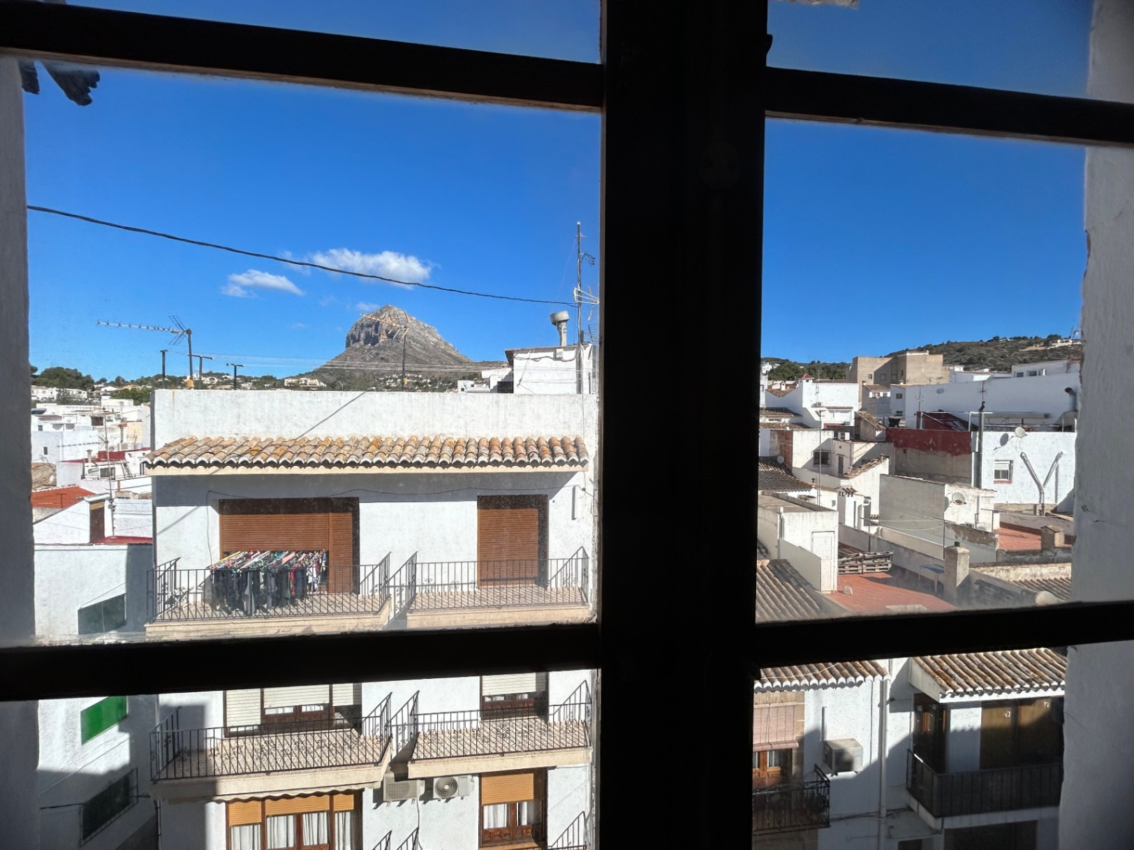 Stadtblick von innen durch einen Fensterrahmen auf die Dächer der Altstadt von Jávea und den Gipfel des Montgó-Massivs bei klarem Himmel.