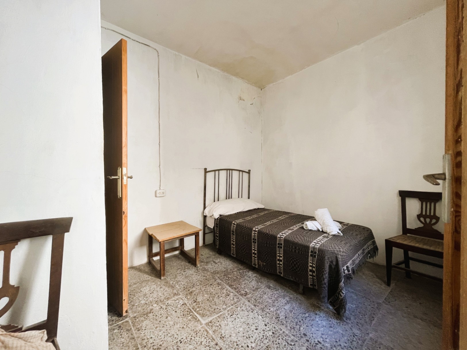 Schlichtes Schlafzimmer mit originalem Terrazzoboden, Einzelbett mit Metallkopfteil und traditionellen Holzmöbeln in einem Raum mit weißen Wänden.
