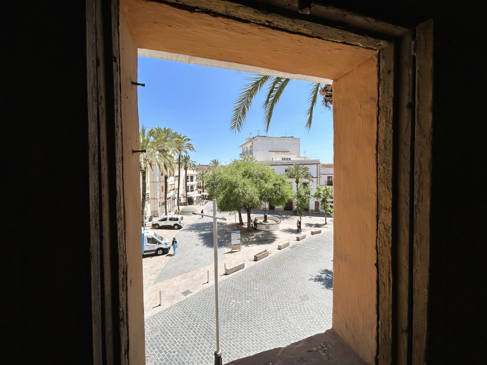 Blick aus dem Innenraum auf einen gepflasterten Platz in der Altstadt, der die traditionellen dicken Mauern und die sonnige Umgebung mit Palmen zeigt.