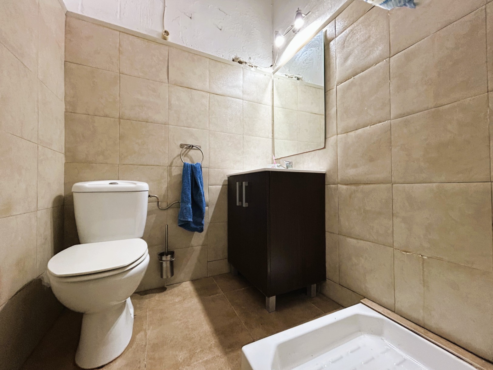 Funktionales Badezimmer mit beigen Wandfliesen, weißem Keramik-WC und dunklem Waschtisch mit integrierter Spiegelbeleuchtung.