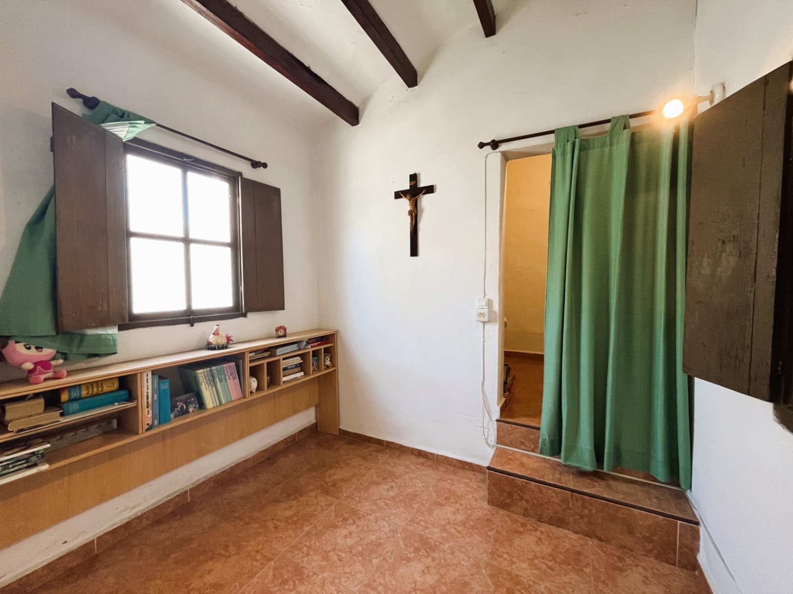 Innenraum mit dunklen Sichtbalken, Terrakottaboden und einem Regal unter dem Holzfenster. Ein erhöhter Durchgang mit Stufen und Vorhang trennt die Wohnbereiche.