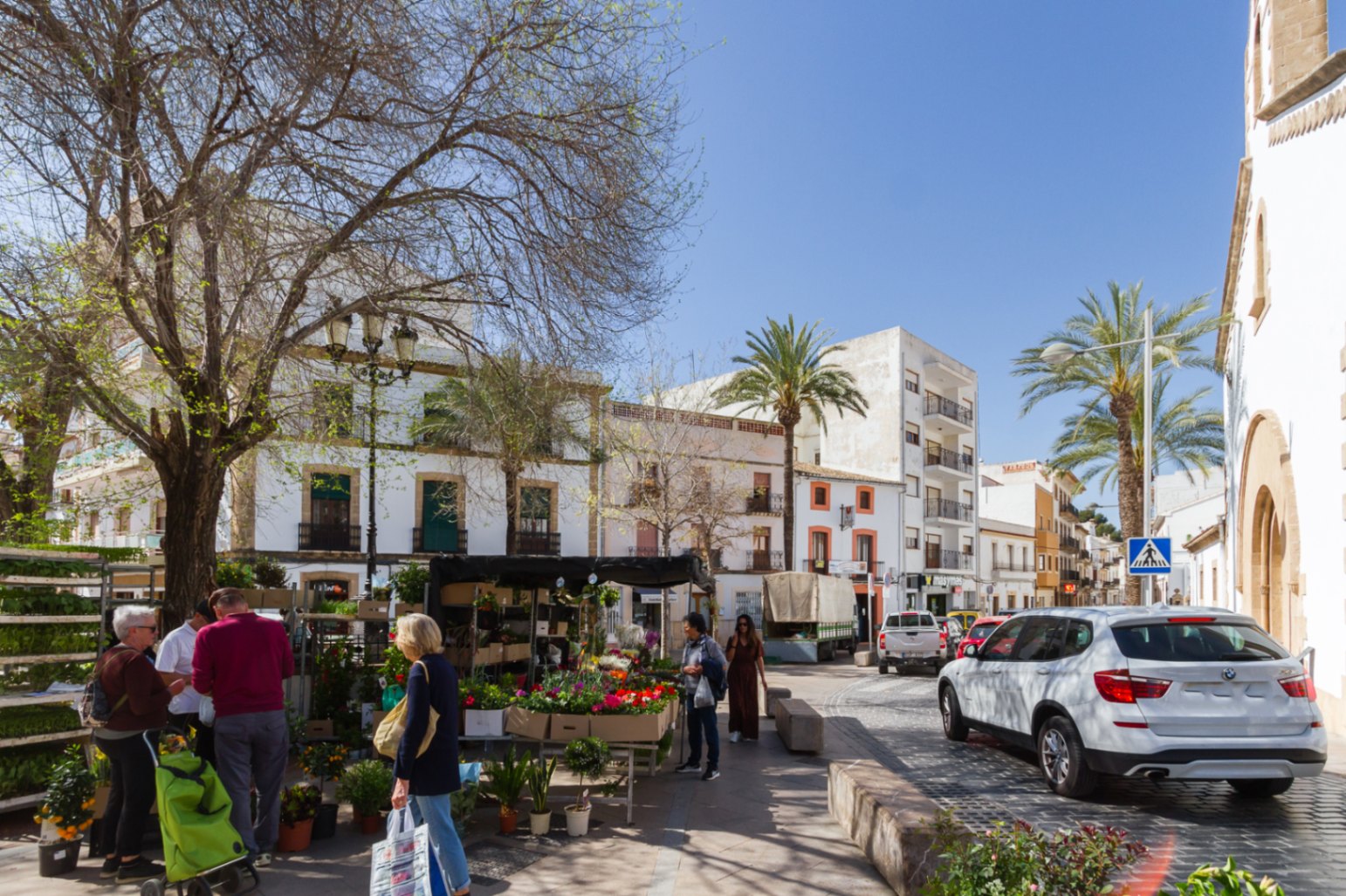 Platz in der Altstadt von Jávea mit einem Blumenstand, umgeben von traditioneller weißer Architektur, Kopfsteinpflaster und Palmen bei hellem Tageslicht.