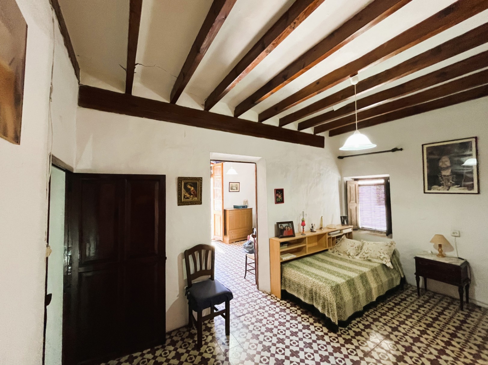 Schlafzimmer mit dunklen Holzbalkendecken, originalen hydraulischen Bodenfliesen und weißen Wänden. Es verfügt über ein kleines Fenster mit Fensterläden und Einbauregale.