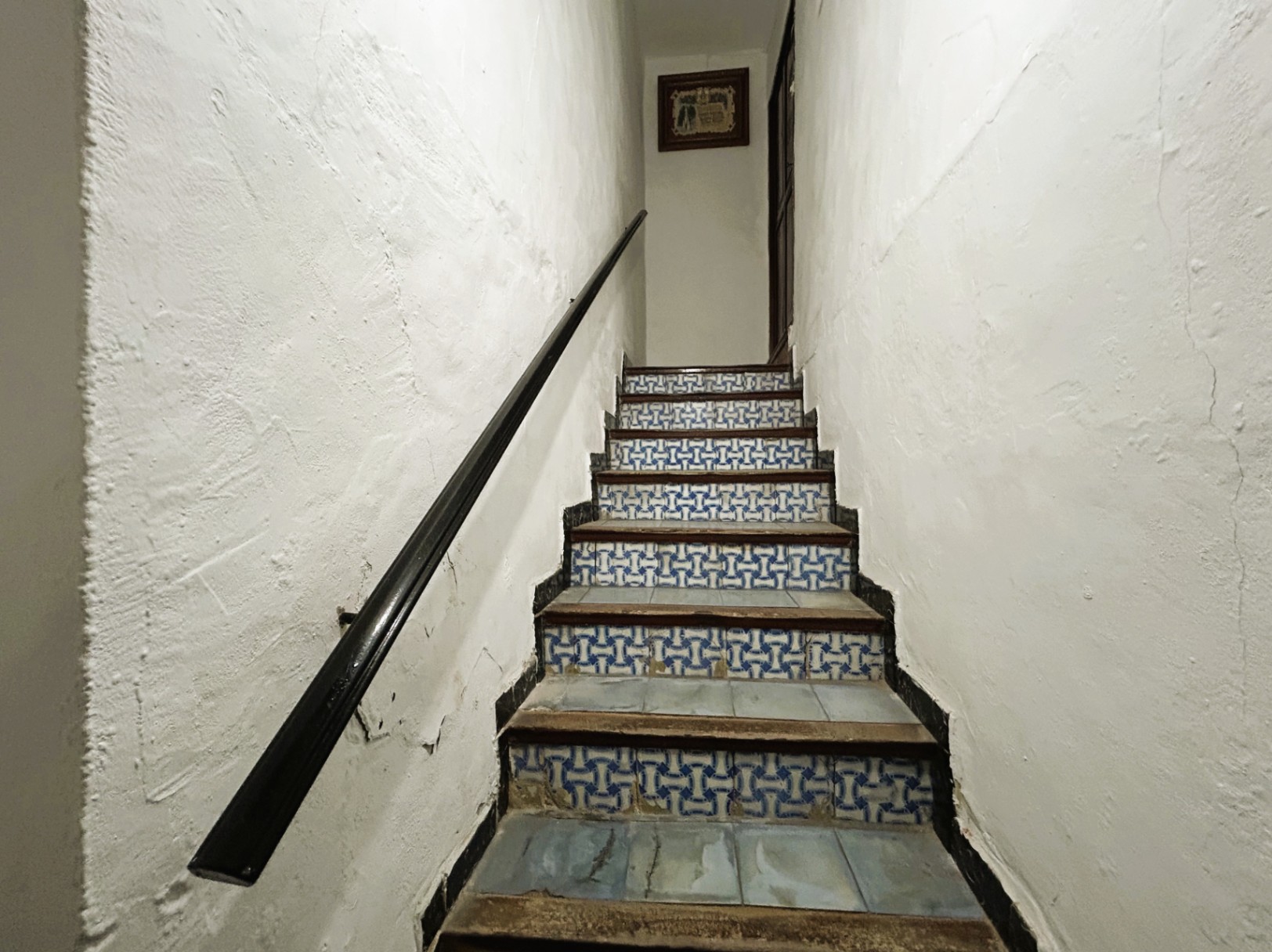 Traditionelle Treppe mit blau-weiß gemusterten Setzstufen aus Fliesen, Holztritten und einem dunklen Handlauf an weiß verputzten Wänden.