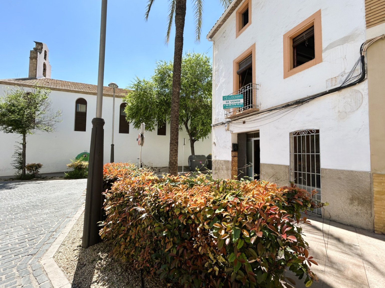 Traditionelle weiße Fassade mit ockerfarbenen Fensterrahmen an einem gepflasterten Platz neben einer historischen Kirche in der Altstadt von Jávea.