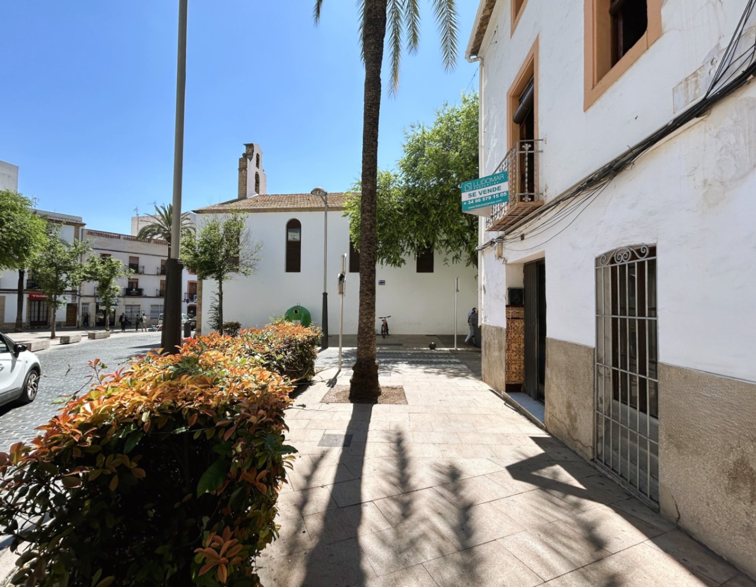 Weiße Fassade mit Steinsockel in der Altstadt von Jávea, direkt an einem Fußgängerplatz mit Palmen und Blick auf einen historischen Glockenturm gelegen.