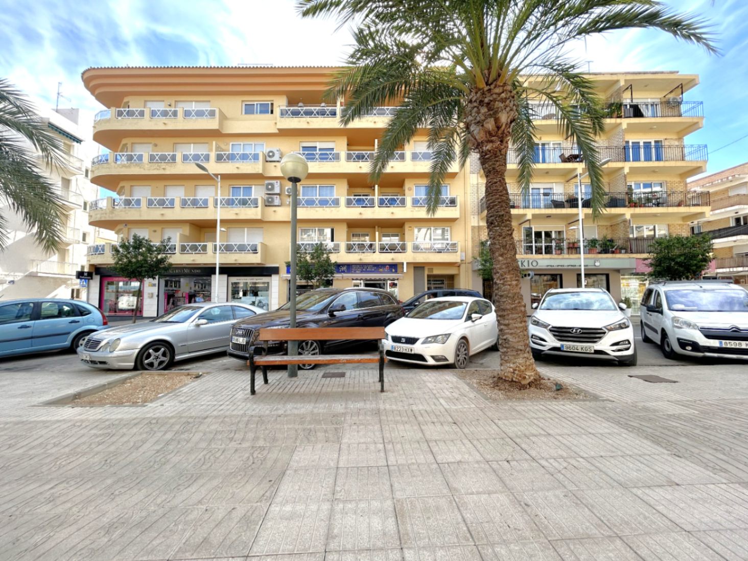 Außenansicht des Gebäudes im Hafen von Jávea mit ockerfarbener Fassade und Balkonen mit weißen Geländern an einem Platz mit Palmen.