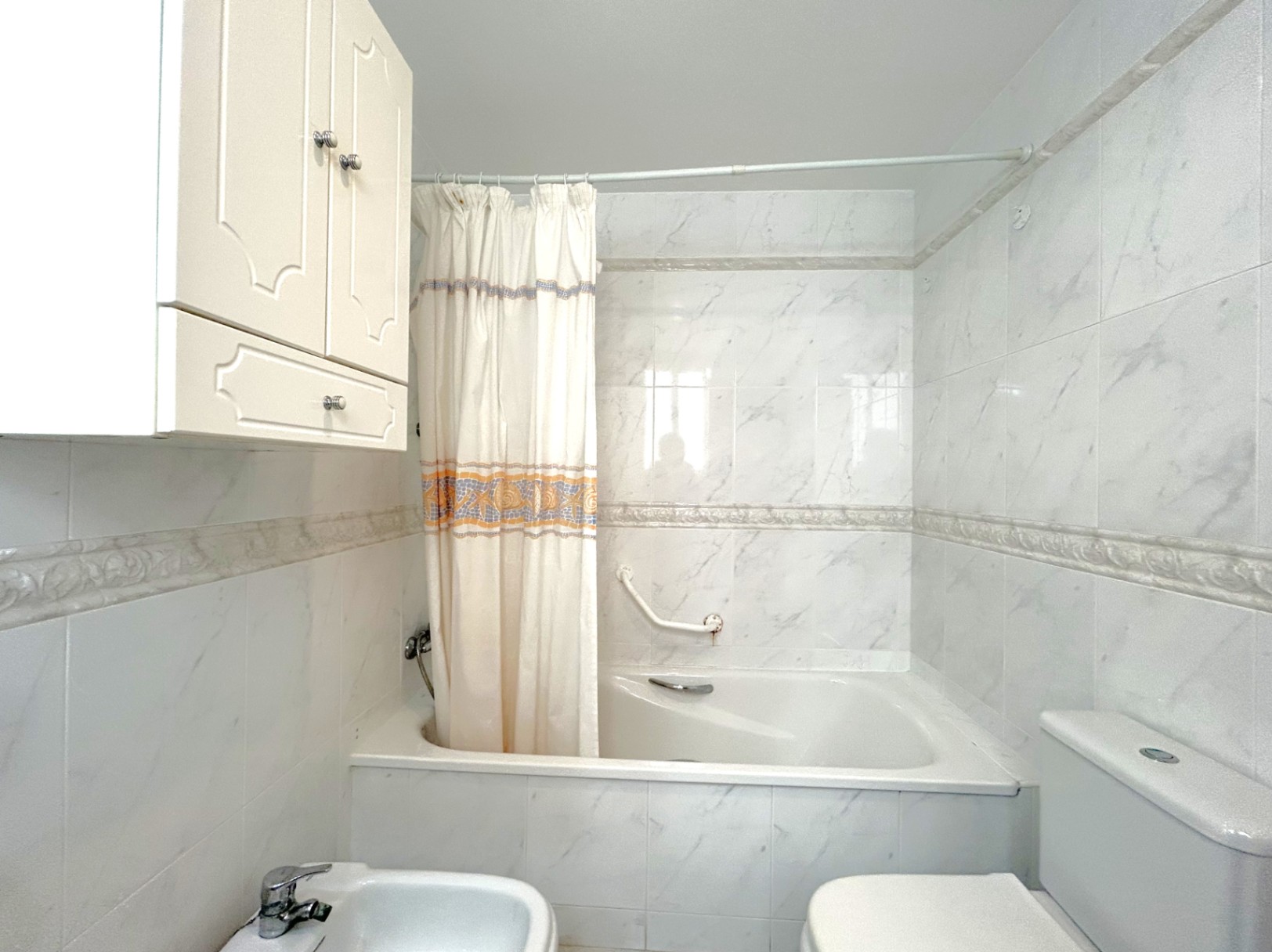 Badezimmer mit Einbaubadewanne, Fliesen in Marmoroptik und dekorativer Bordüre. Ausgestattet mit Bidet, WC und einem weißen Hängeschrank für Stauraum.