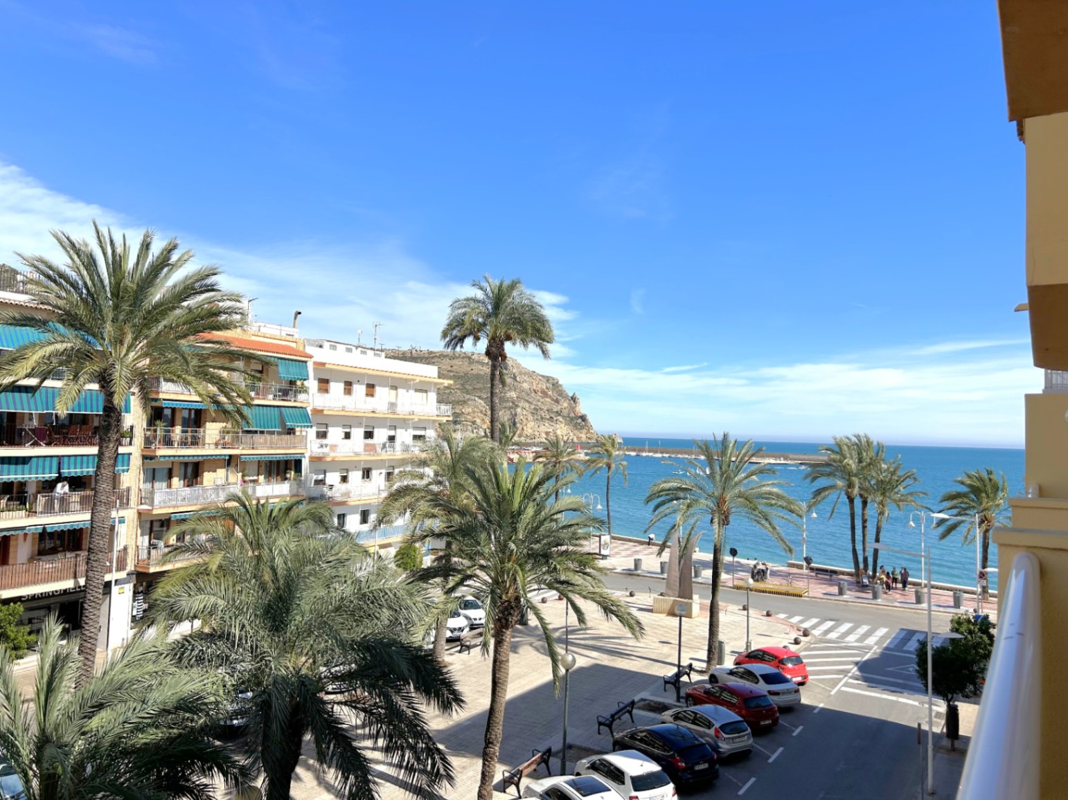Blick vom Balkon auf die Promenade von Puerto de Jávea mit Palmen, dem Mittelmeer und den markanten Küstenfelsen unter strahlend blauem Himmel.