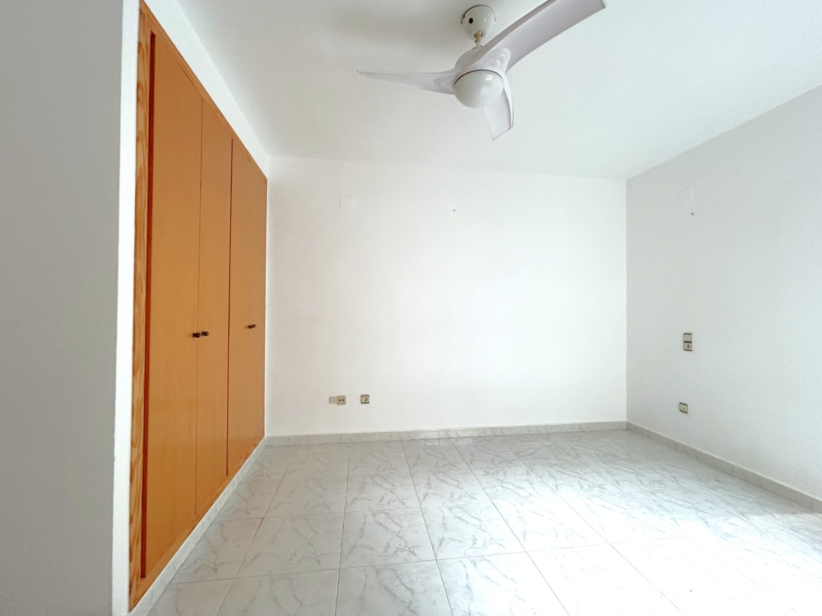 Helles Schlafzimmer mit großem Einbauschrank aus Holz, Bodenfliesen in Marmoroptik und weißem Deckenventilator.