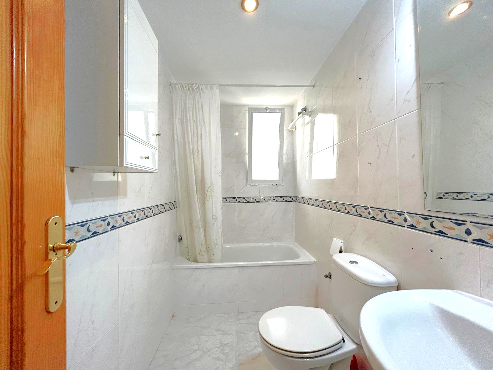 Komplettes Badezimmer mit Badewanne, Marmoreffekt-Fliesen und dekorativer Bordüre. Ein Fenster sorgt für natürliches Licht und eine helle Atmosphäre.