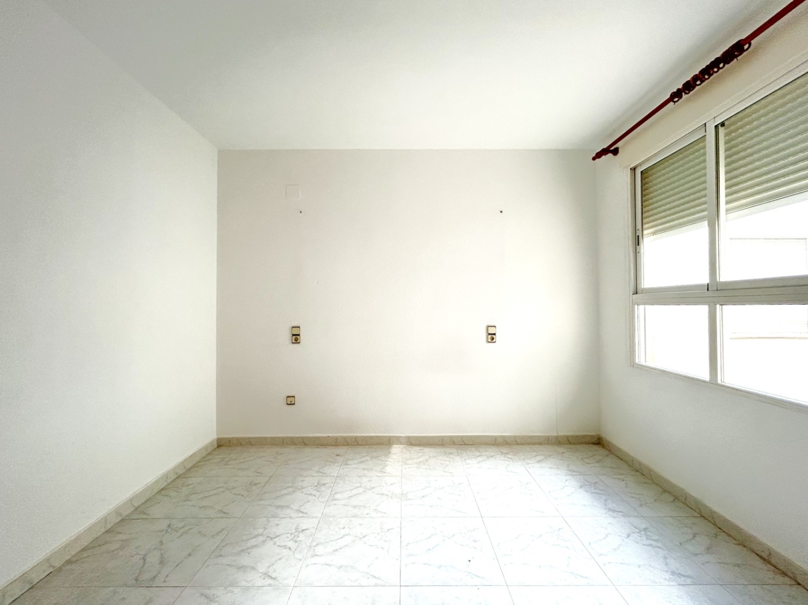 Leeres Schlafzimmer mit hellen Fliesen in Marmoroptik, weißen Wänden und einem Fenster mit Rollladen. Symmetrische Steckdosen sind für den Bettbereich vorinstalliert.