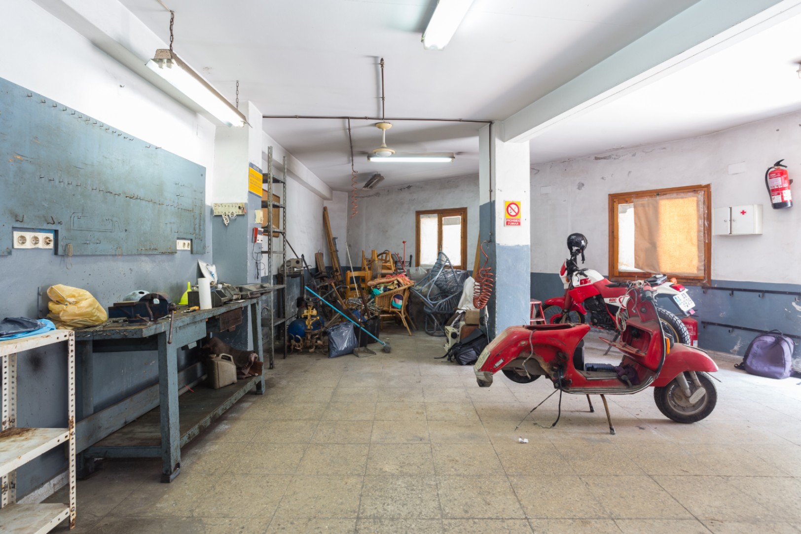 Geräumige Werkstatt oder Garagenbereich mit Werkbank, Metallregalen und Fliesenboden, ausgestattet mit Leuchtstoffröhren und Deckenventilatoren.