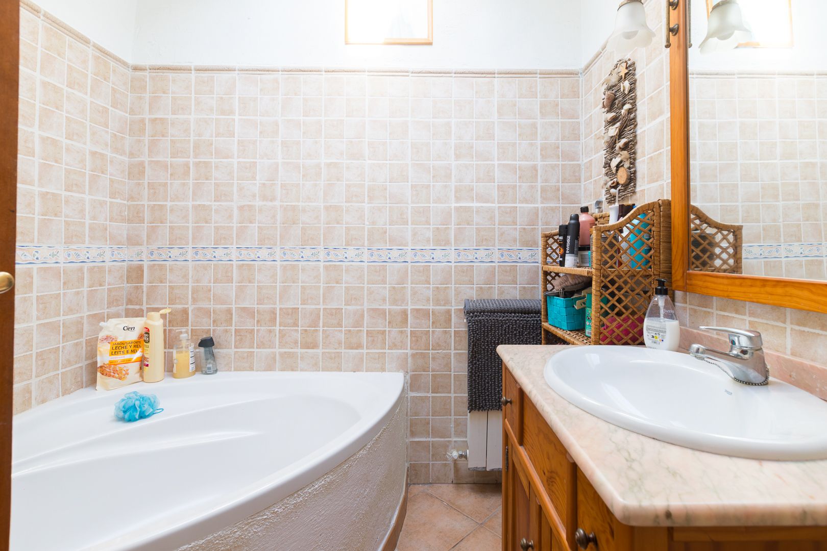 Badezimmer mit Eckbadewanne, Waschtisch aus Holz mit Marmorplatte und beigefarbenen Wandfliesen mit dekorativer Bordüre.