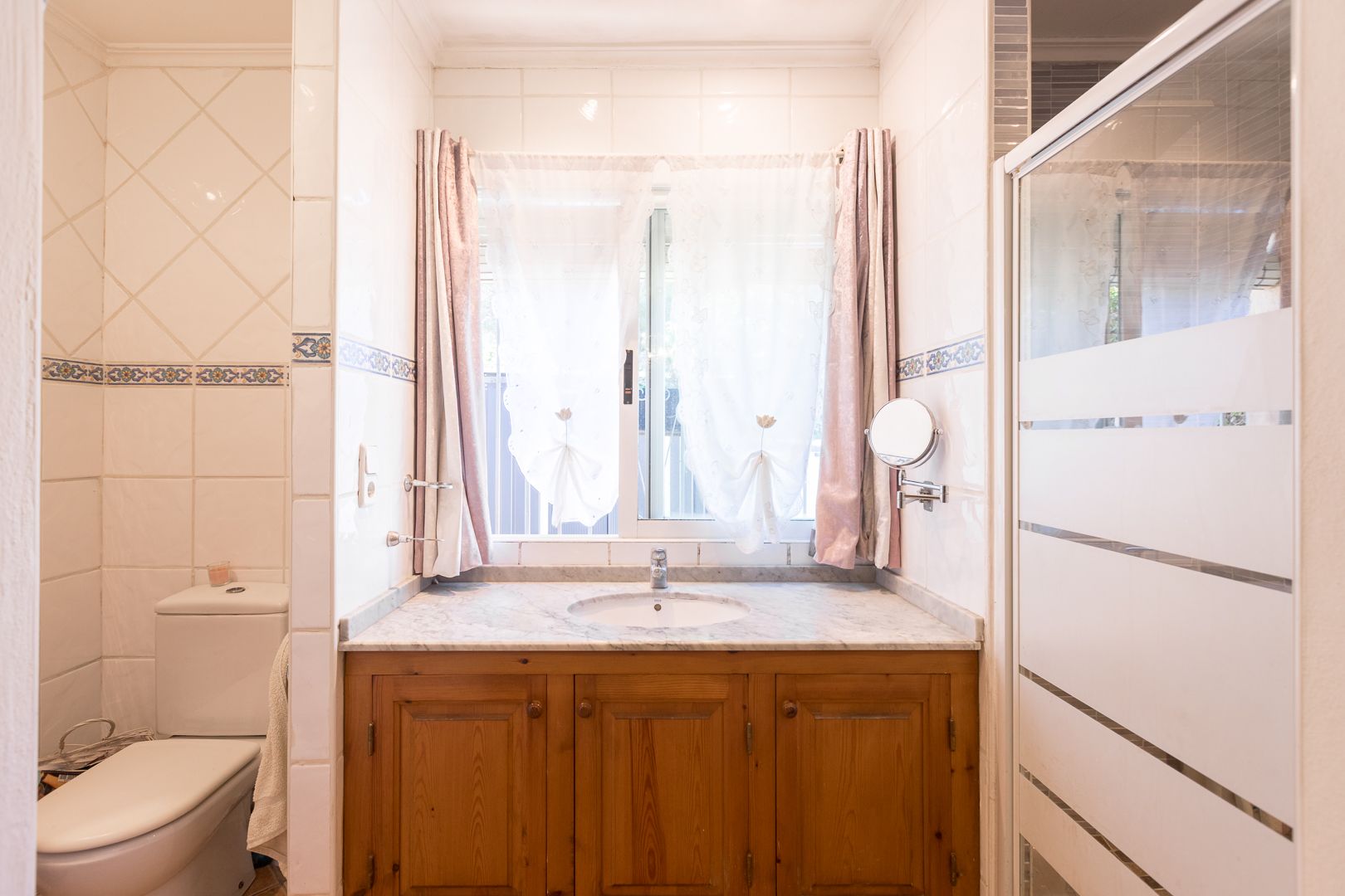 Badezimmer mit Holzwaschtisch, Marmorplatte und Fenster. Ausgestattet mit einer Glasduschwand mit Sichtschutzstreifen und dekorativen Wandfliesen.