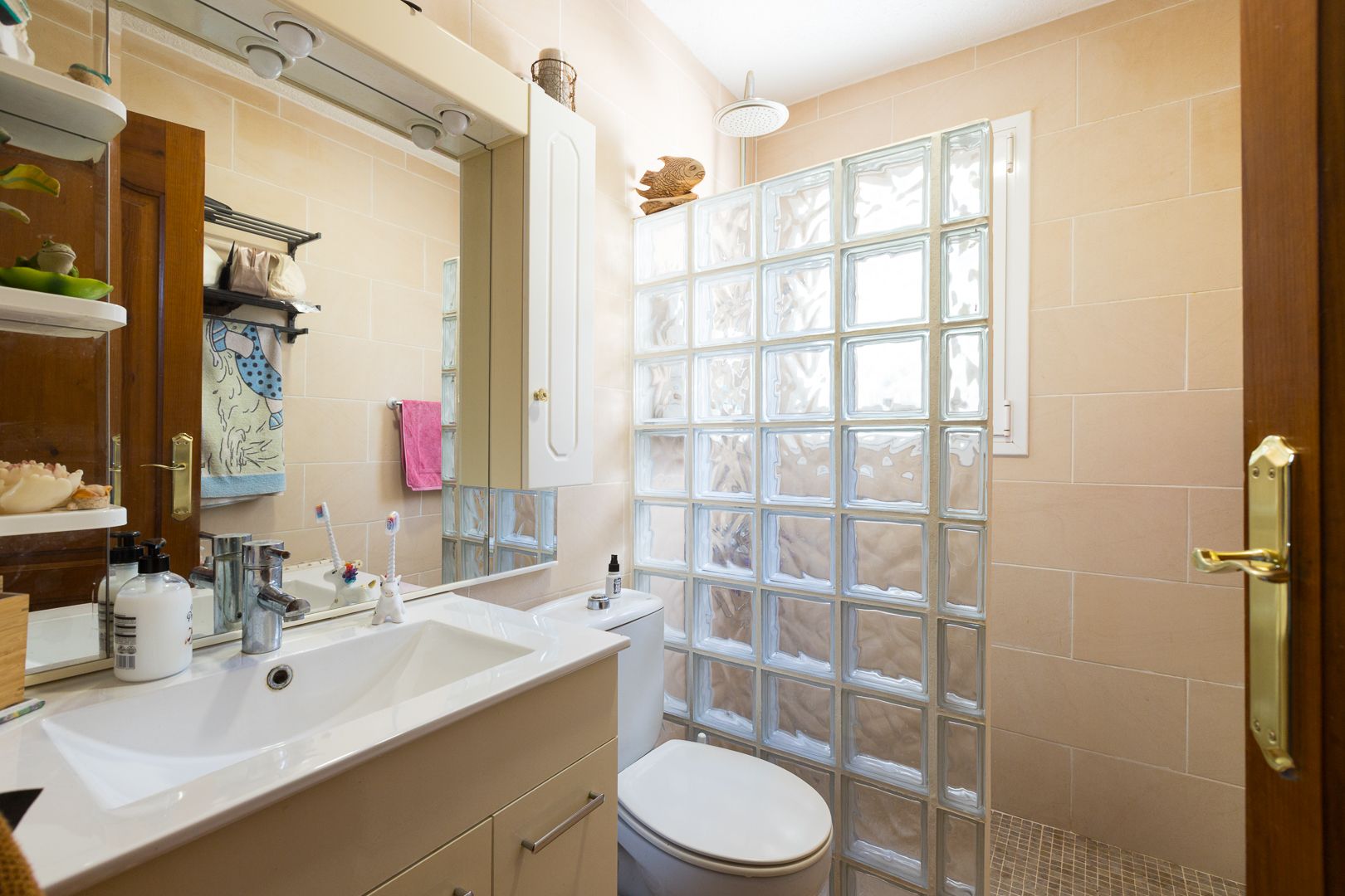 Funktionales Badezimmer mit modernem Waschtisch, großem Spiegel und einer Dusche, die durch eine lichtdurchlässige Glasbausteinwand vom restlichen Raum abgetrennt ist.