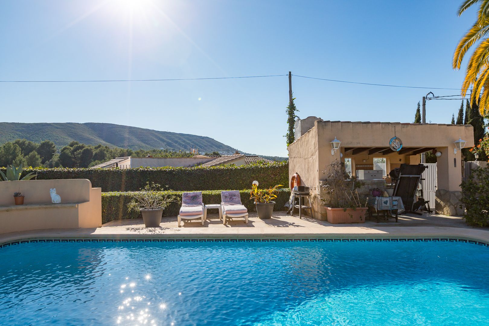 Gefliester Pool mit Sonnenterrasse, Liegestühlen und einer überdachten Außenküche, eingebettet in eine Gartenanlage mit Blick auf die nahen Berge.