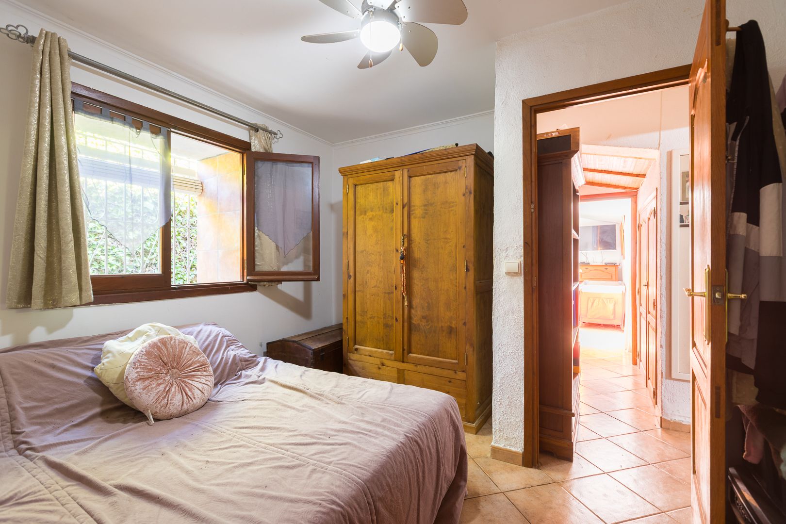 Gemütliches Schlafzimmer mit massivem Holzschrank, Terrakotta-Fliesenboden und Deckenventilator. Ein Holzfenster sorgt für natürliches Licht und Belüftung.