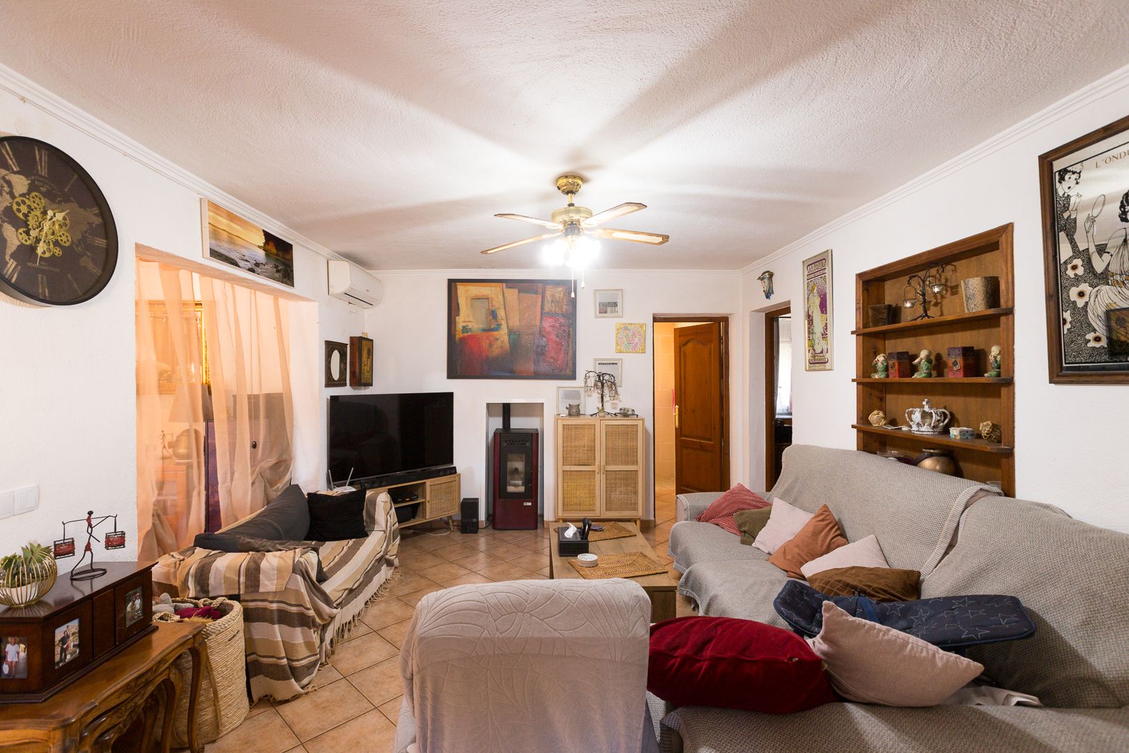 Gemütliches Wohnzimmer mit Terrakotta-Fliesen, Pelletofen, Klimaanlage und Deckenventilator. Ausgestattet mit integrierten Holzregalen und Zugang zu den weiteren Räumen.