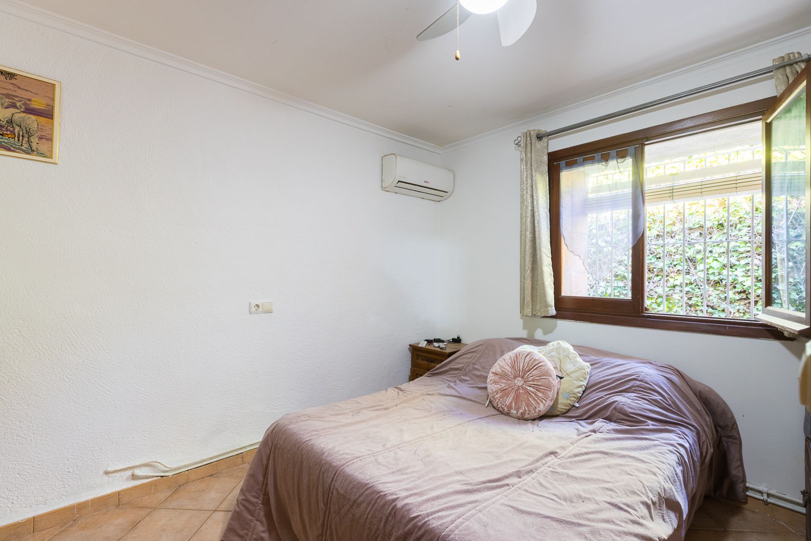 Helles Schlafzimmer mit Split-Klimaanlage, Deckenventilator und Holzfenster mit Blick auf die umliegende Gartenbepflanzung.