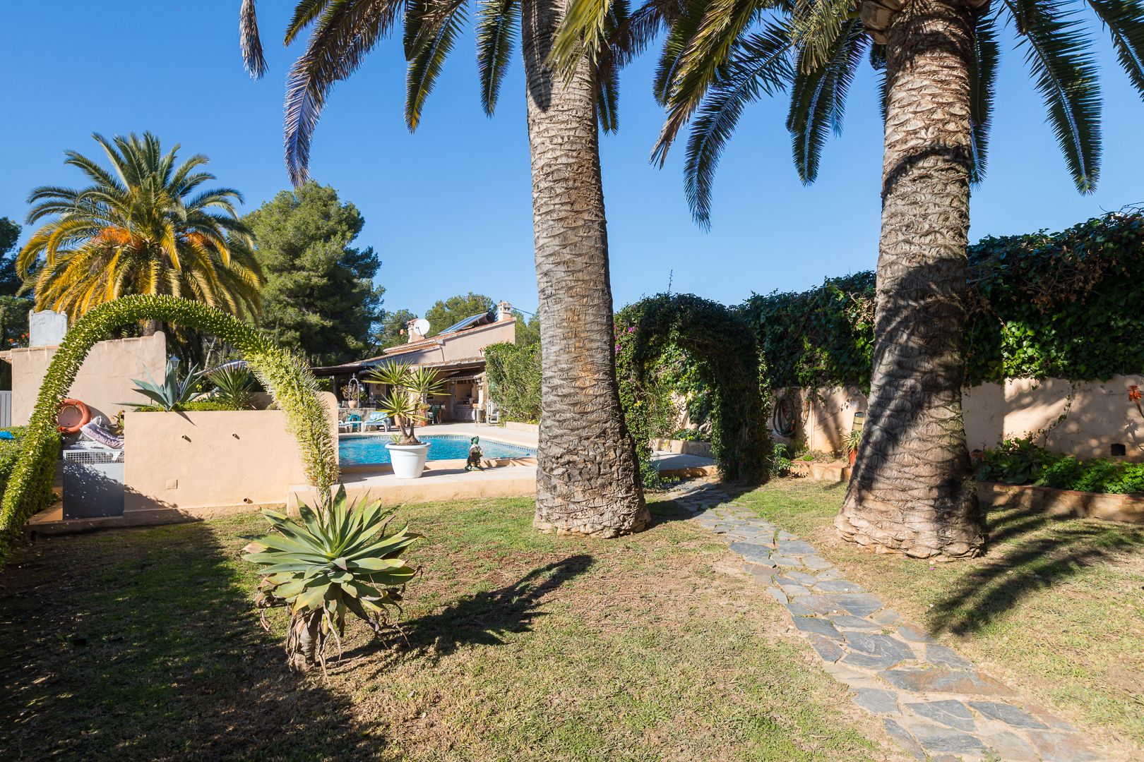 Mediterraner Garten mit hohen Palmen, einem Natursteinweg und Blick auf den Pool sowie die Villa mit Solaranlage.