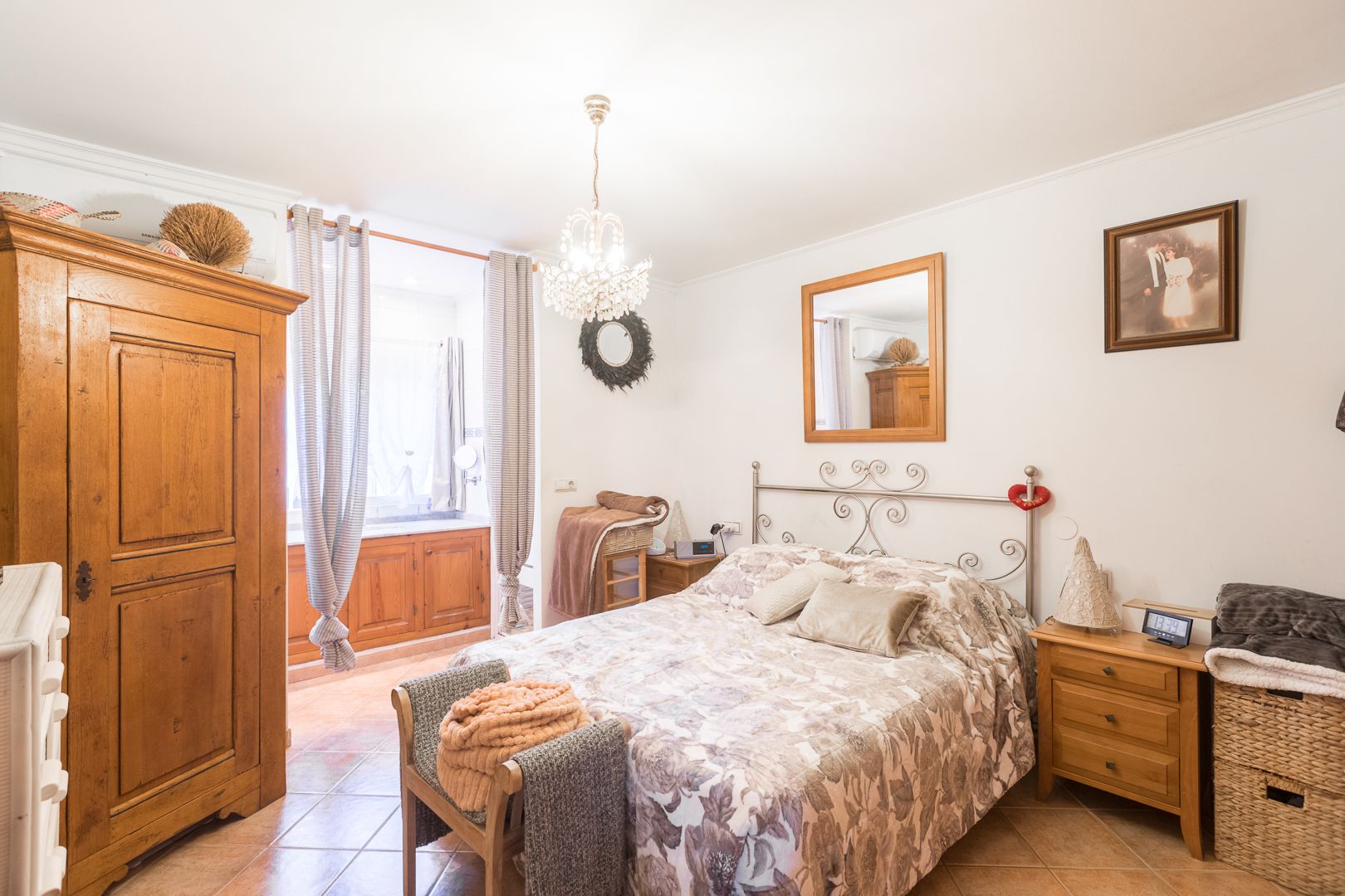 Schlafzimmer mit Keramikfliesen, klassischem Holzschrank und schmiedeeisernem Kopfteil. Ausgestattet mit Klimaanlage, Kristallleuchter und viel Tageslicht.