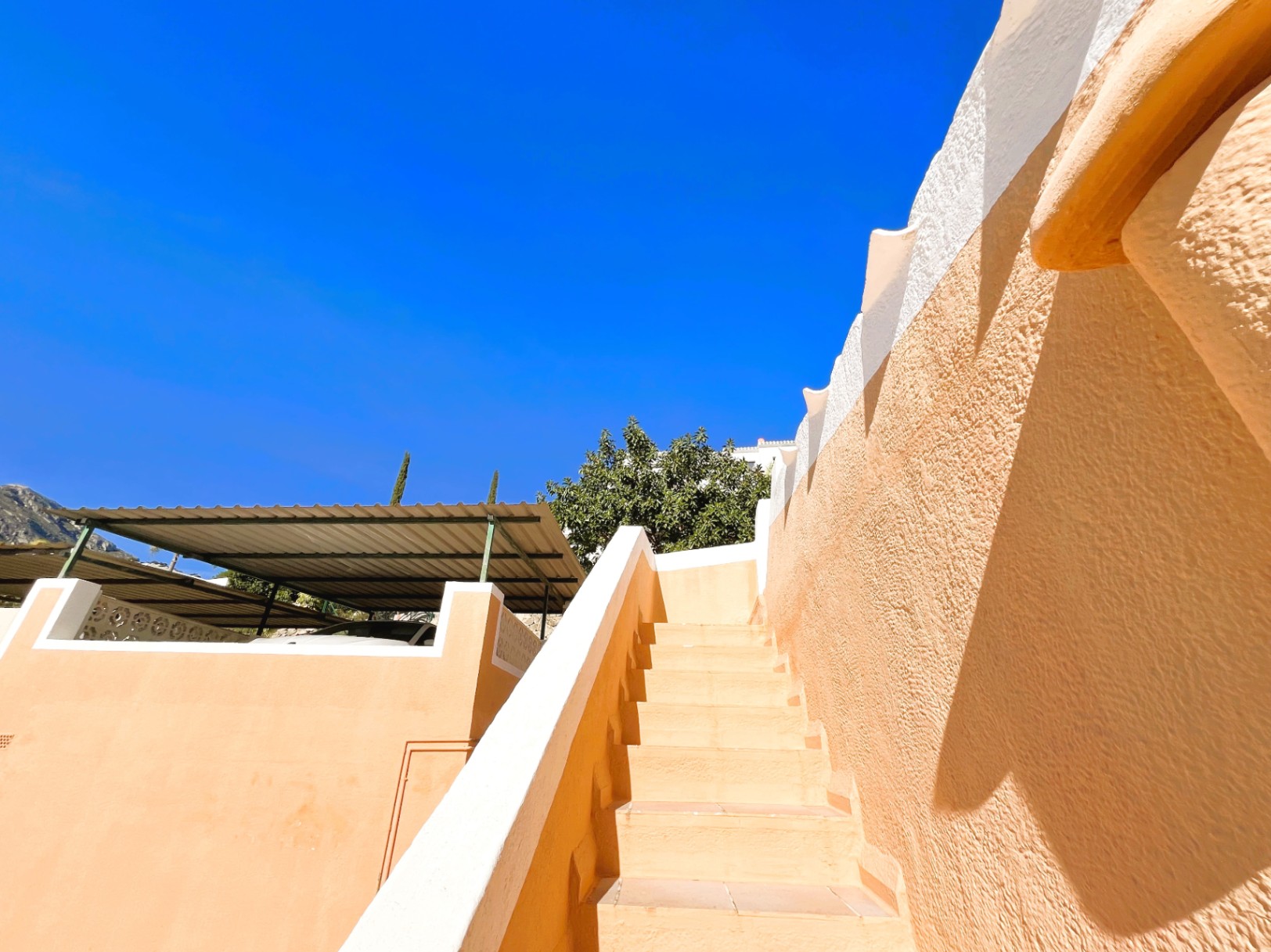 Außenliegende geflieste Treppe mit terrakottafarbenen Wänden und weißem Abschluss, direkt neben dem überdachten Parkplatz mit Metallpergola.