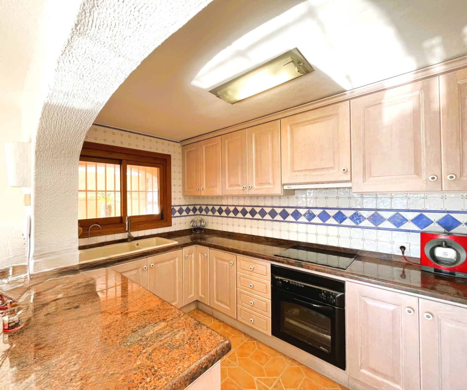 Einbauküche mit hellen Holzfronten, Granitarbeitsplatte und Fliesenspiegel mit blauen Akzenten. Ausgestattet mit integriertem Backofen, Cerankochfeld und Holzfenster.