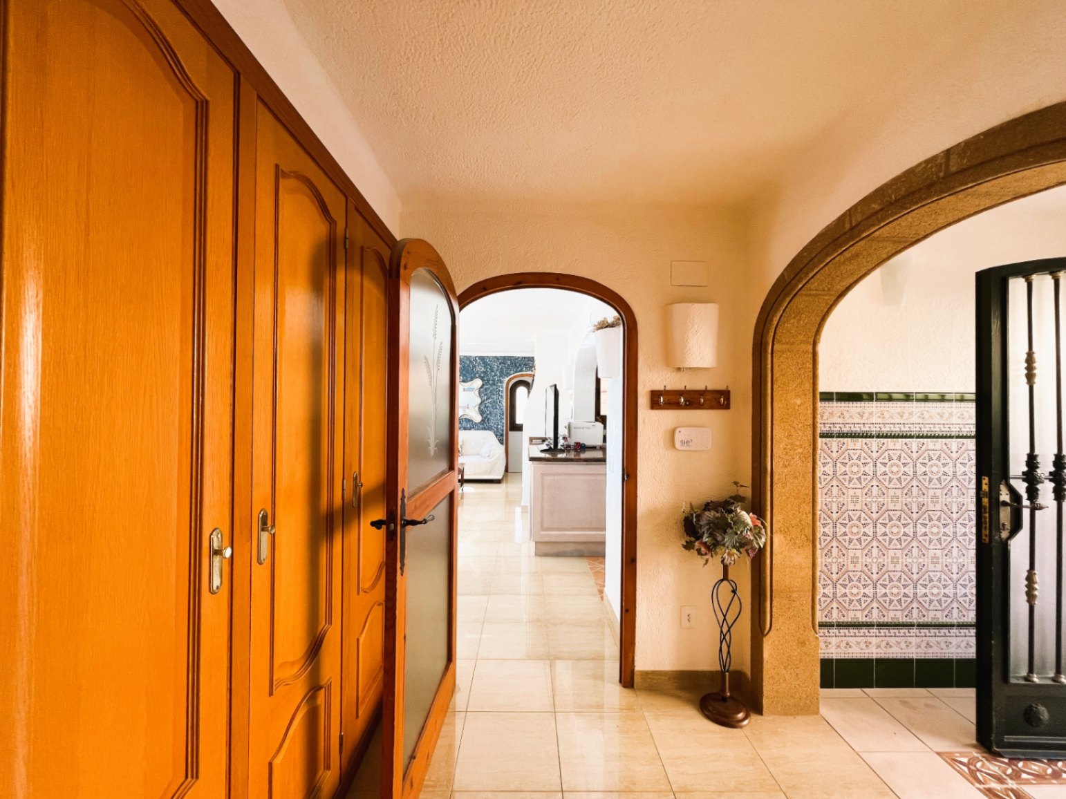 Eingangsbereich mit Einbauschränken aus Holz, glänzenden Fliesenböden und Rundbögen, die zum Wohnzimmer und einem Bereich mit traditionellen Kacheln führen.