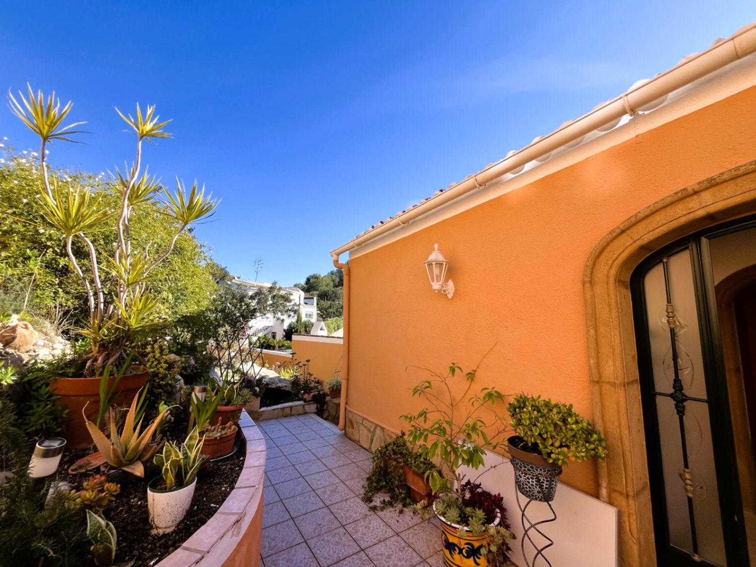 Gefliester Außenweg mit geschwungenem Pflanzkübel, ockerfarbener Fassade mit Natursteinbogen und mediterranen Sukkulenten.