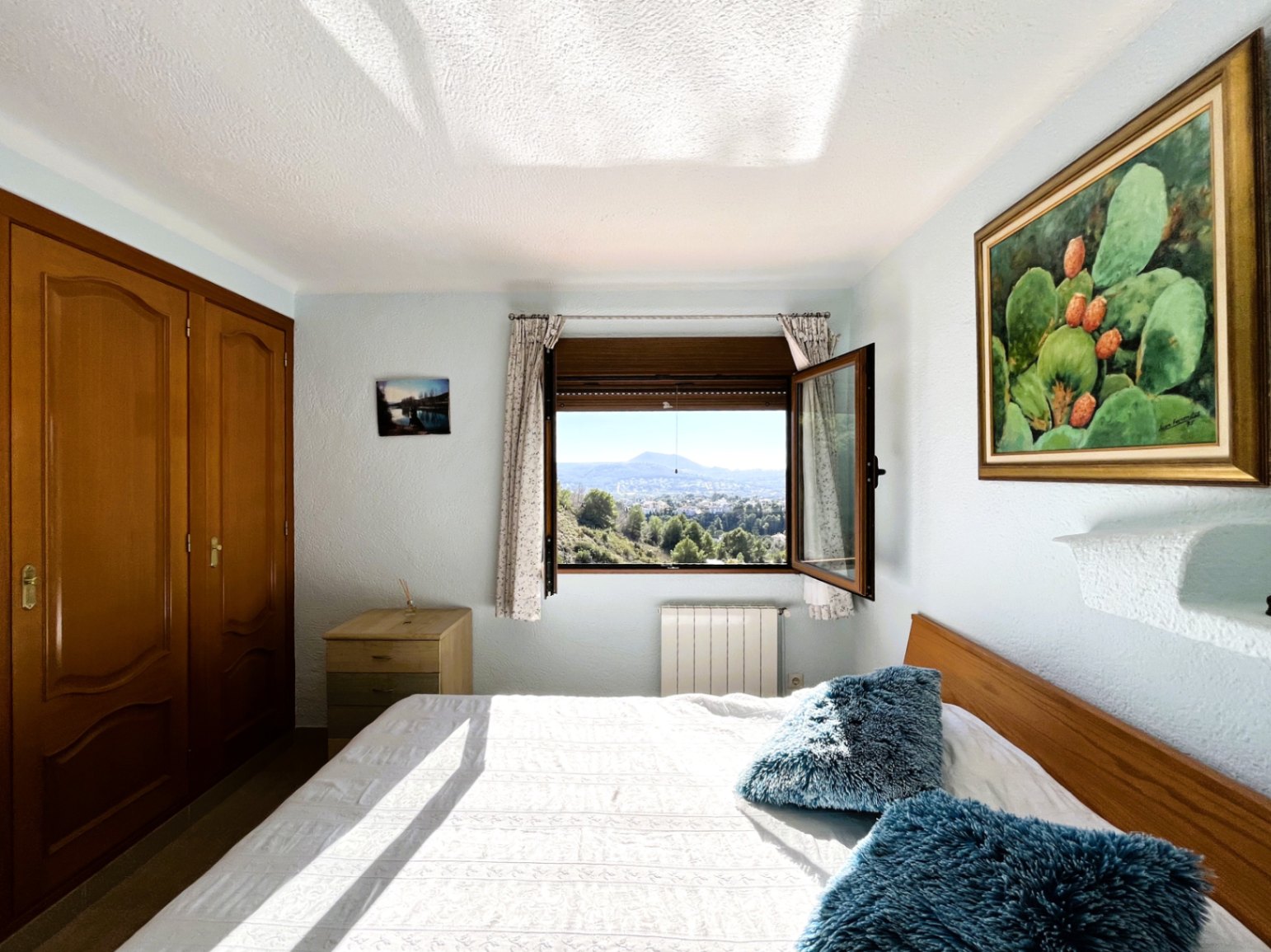 Helles Schlafzimmer mit Einbauschränken aus Holz, Heizkörper und offenem Fenster mit Panoramablick auf die Berge und die Landschaft des Montgó.