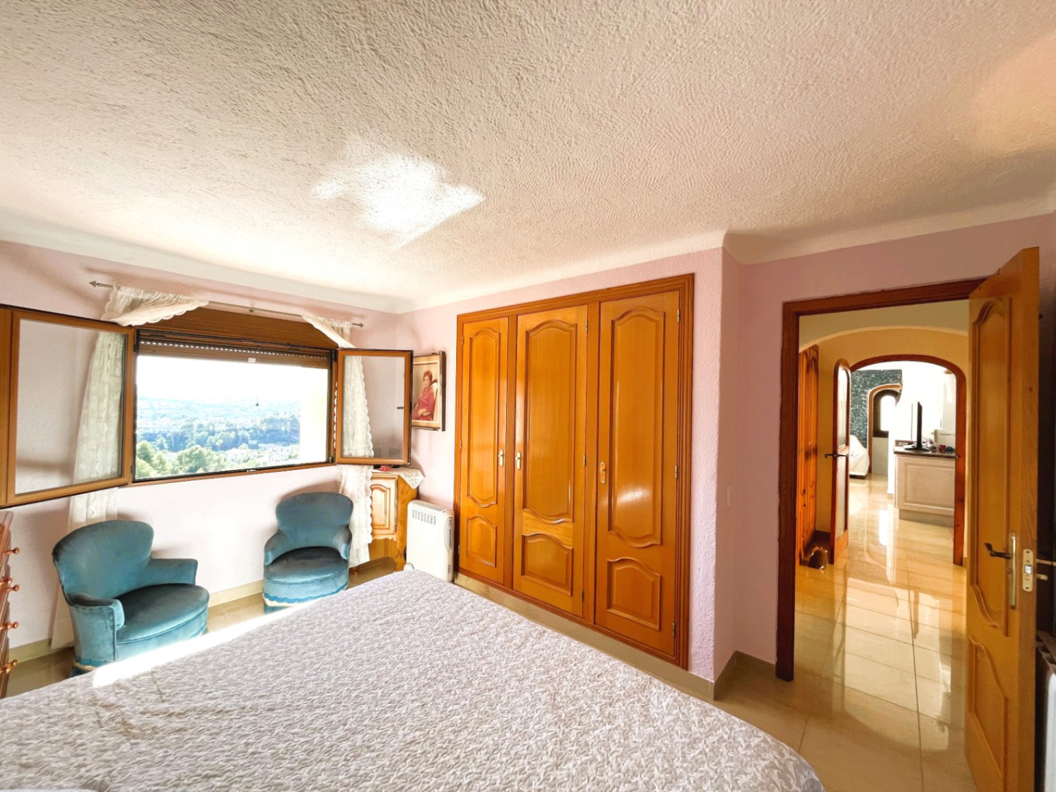 Helles Schlafzimmer mit Einbauschränken aus Holz, Samtsesseln und einem Fenster mit weitem Panoramablick auf die umliegende Landschaft.