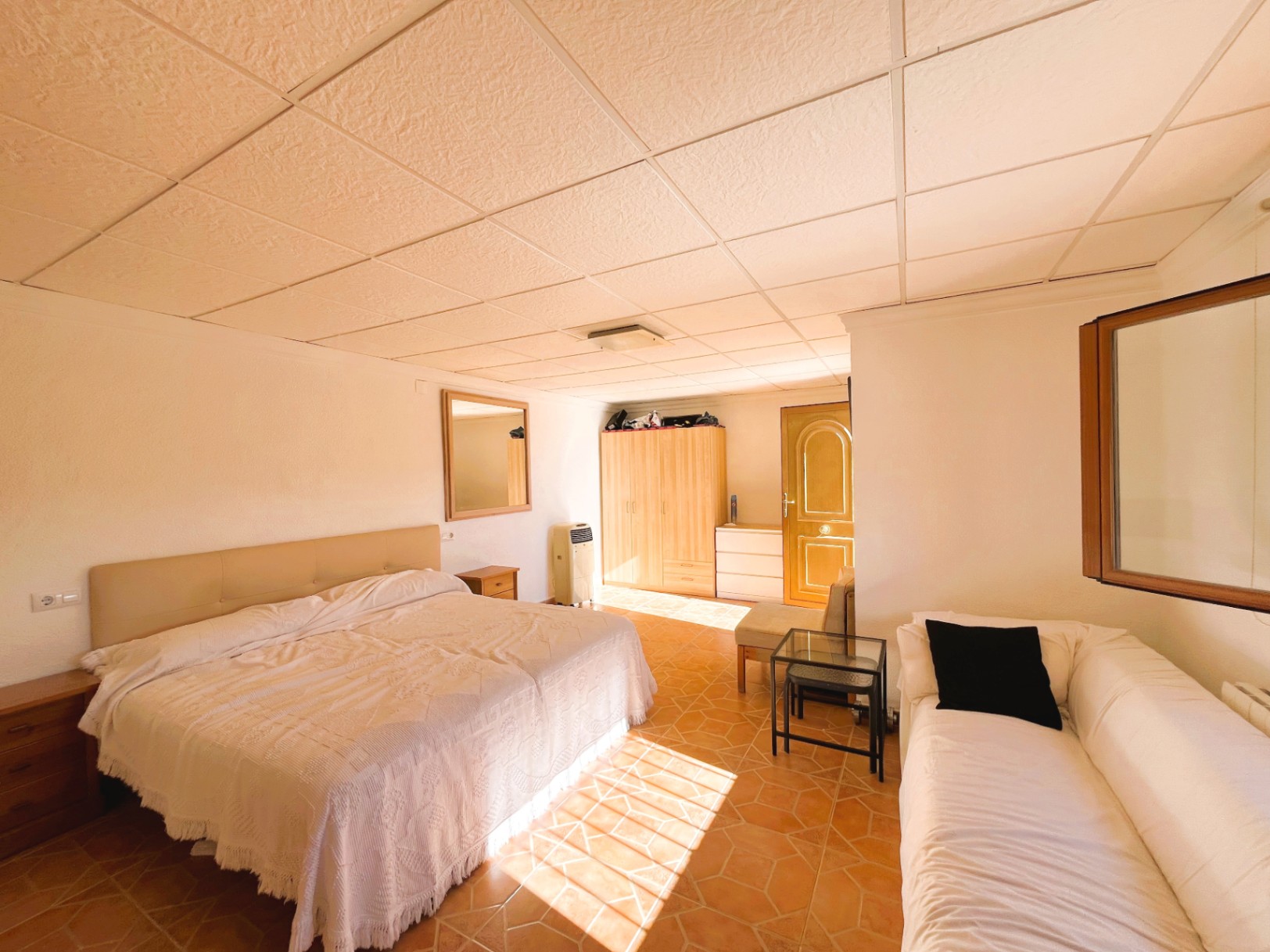Schlafzimmer mit Terrakotta-Fliesen, Rasterdecke und integrierter Sitzecke. Natürliches Licht betont die Holzmöbel und die funktionale Raumaufteilung.